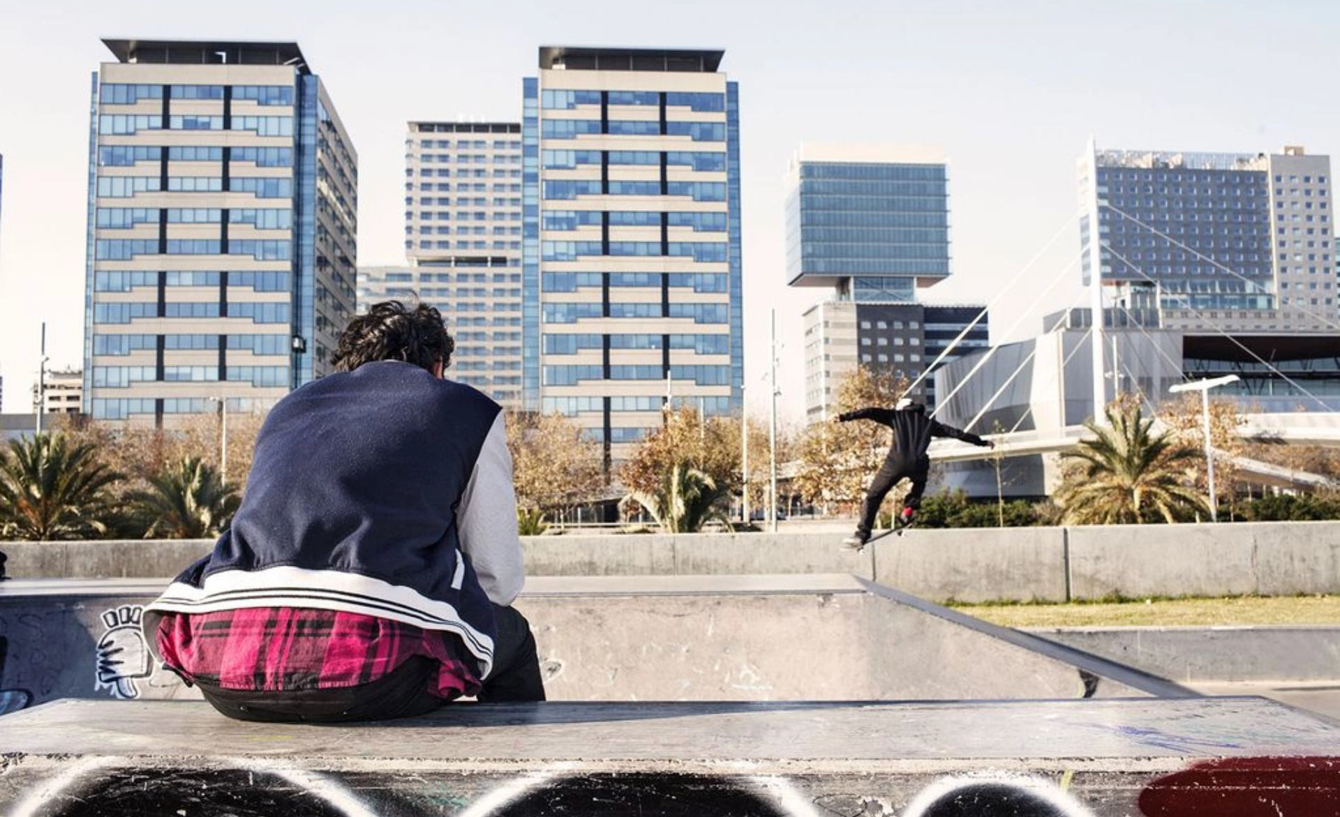 Dos joves en un skate parc, amb edificis al fons. Un assegut d'esquenes i al fons un altre fent una pirueta.