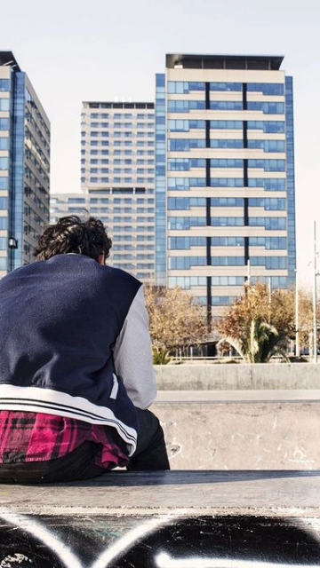 Dos joves en un skate parc, amb edificis al fons. Un assegut d'esquenes i al fons un altre fent una pirueta.